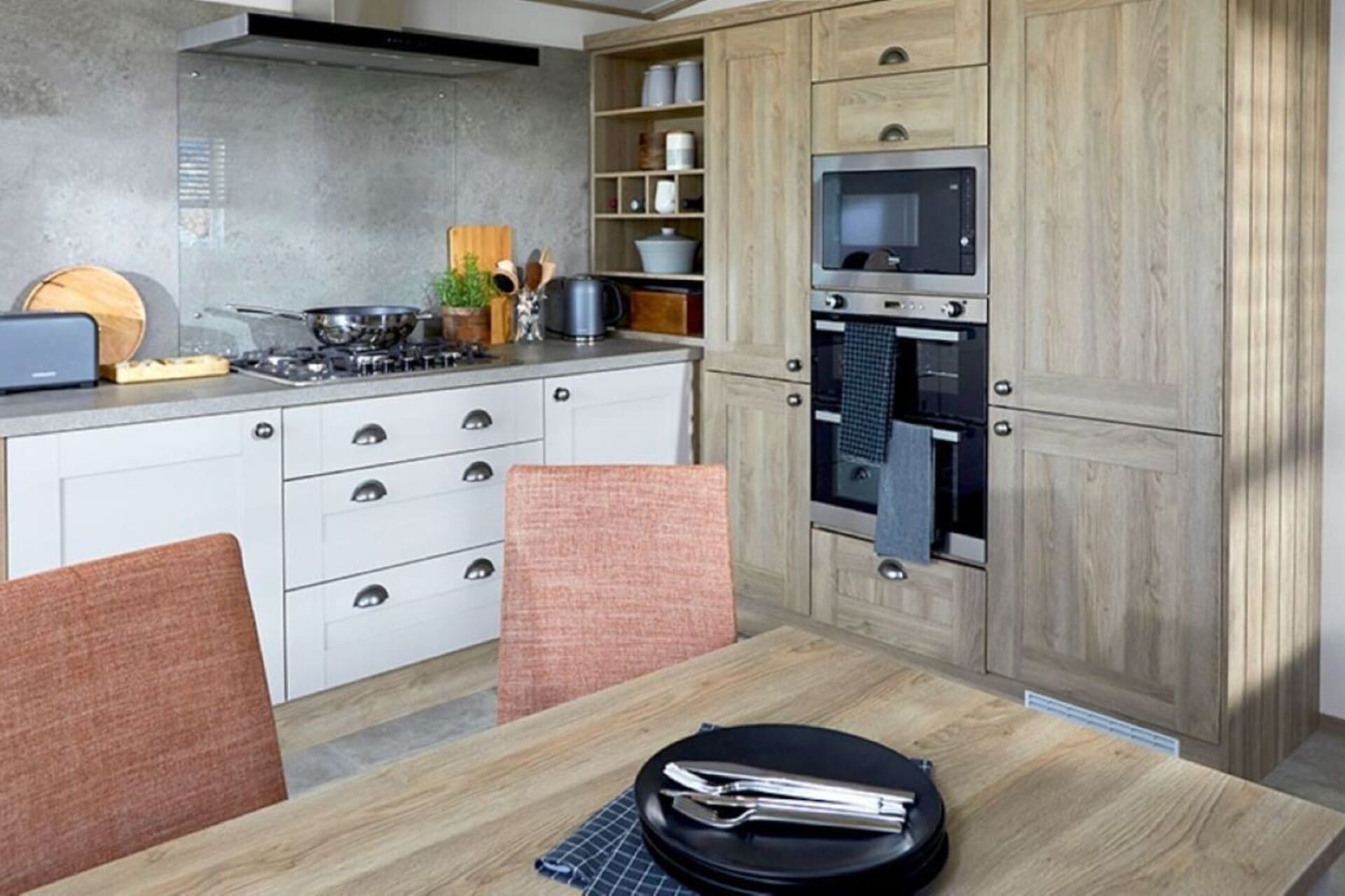 A modern kitchen with white cabinets, a built-in oven, and a dining table with a black plate and tongs.