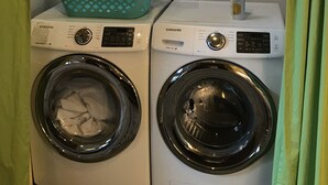 Two front-loading washing machines with control panels, a teal laundry basket on top of the left machine, and a shelf above containing various items like bottles and a container. - Great Monthly Furnished Apartment in Downtown LR (Little Rock)