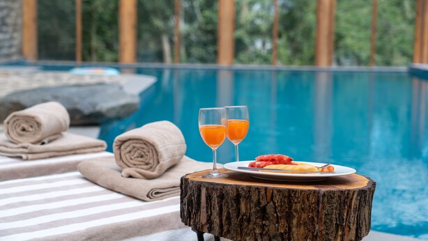 Escena junto a la piscina con una mesa de madera, dos vasos de zumo de naranja, un plato de fruta y toallas.