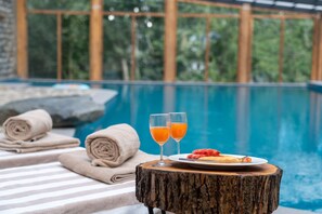 Escena junto a la piscina con una mesa de madera, dos vasos de zumo de naranja, un plato de fruta y toallas.