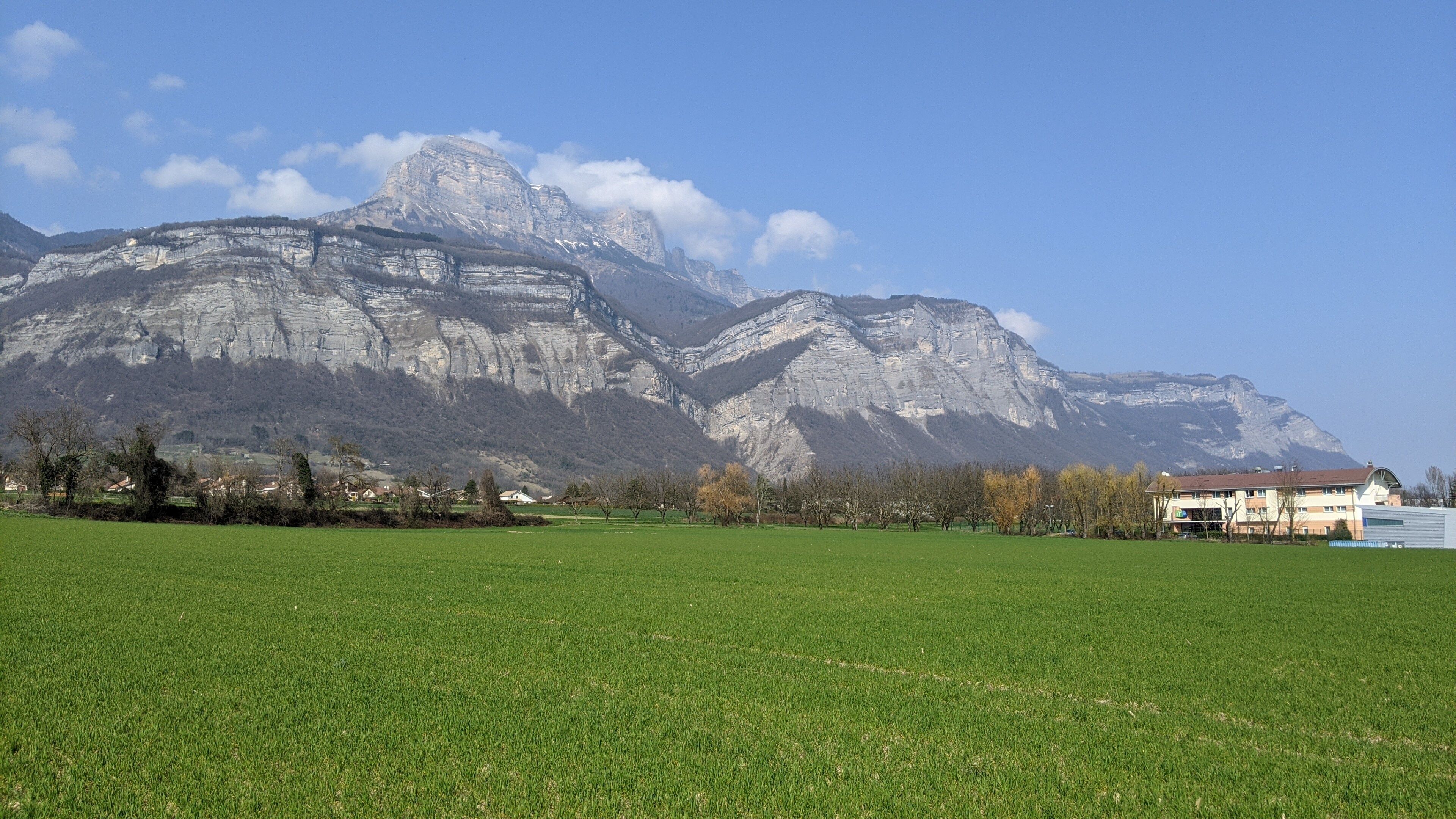 Eine Berglandschaft mit einem grünen Feld im Vordergrund und einem Gebäude mit rotem Dach im Hintergrund.