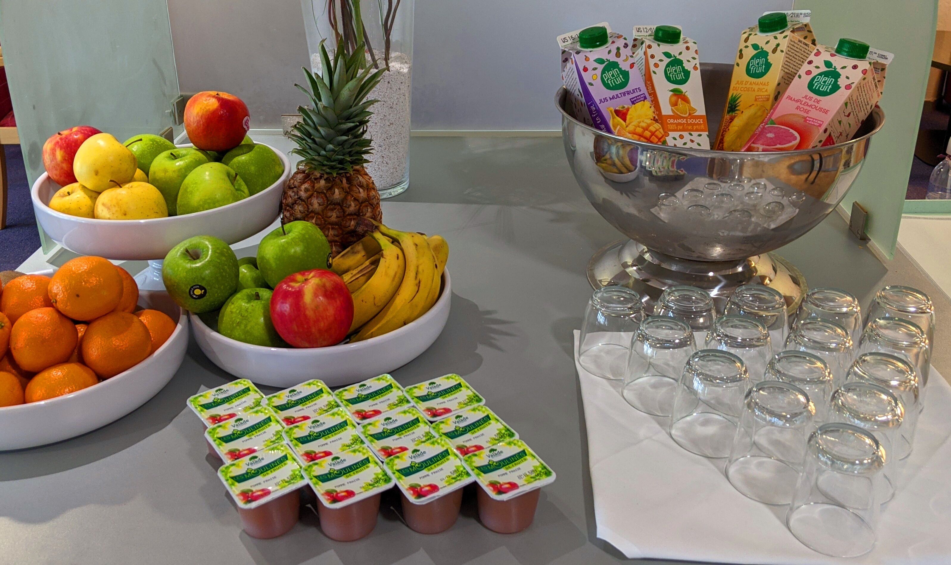 Une table de buffet avec des saladiers remplis de fruits, des briques de jus de fruits et des pots de yaourt.