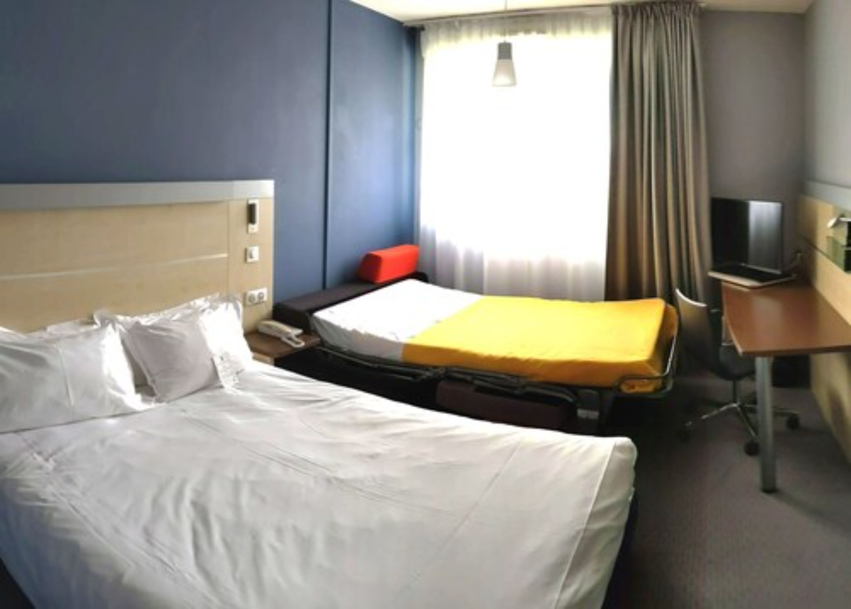 A hotel room with a large bed, a smaller bed, a desk, a chair, a TV, and a window with curtains.