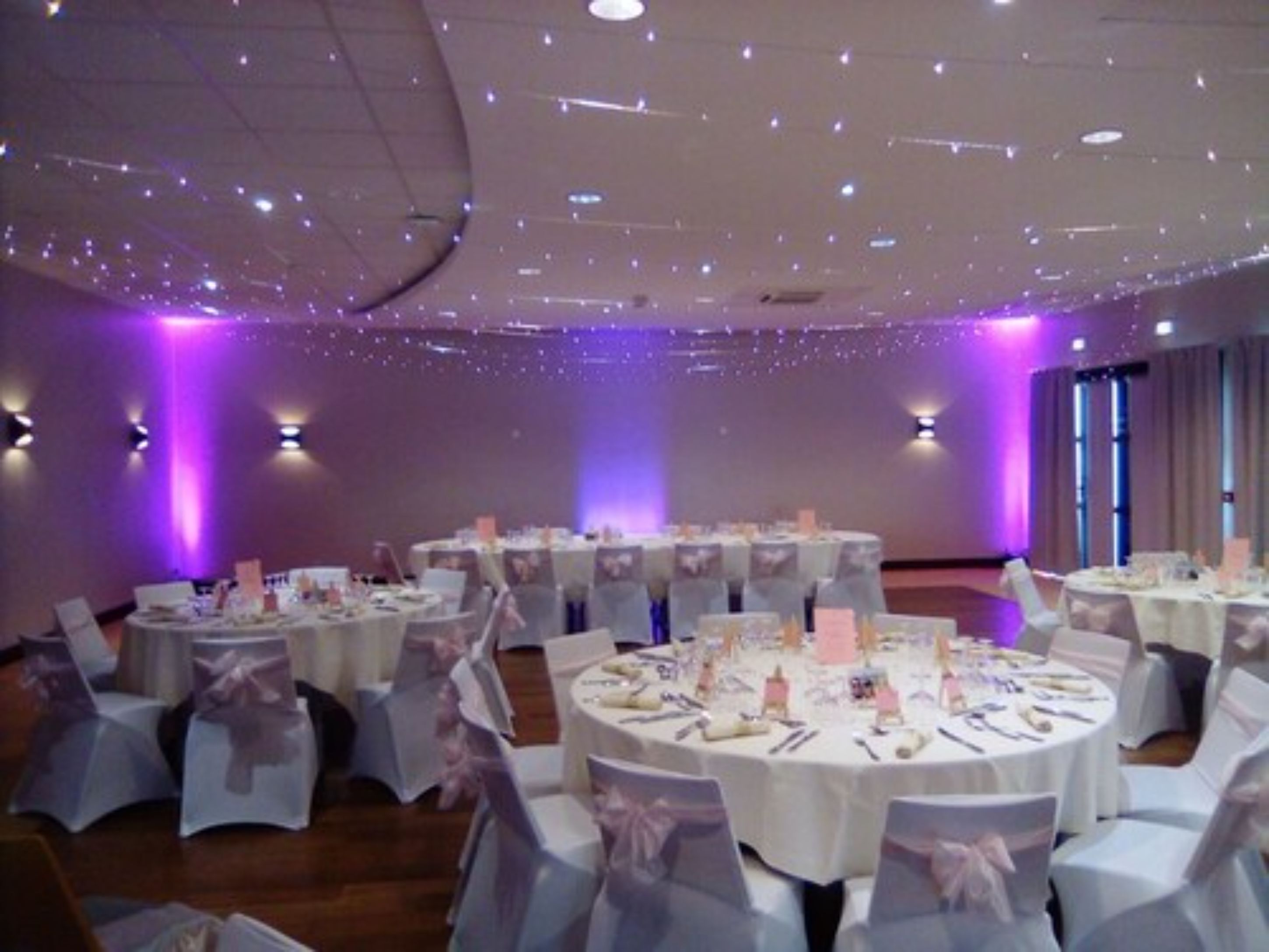 Une salle de banquet avec des tables rondes, des nappes blanches et des chaises recouvertes d’housse blanche et ornées de rubans roses.