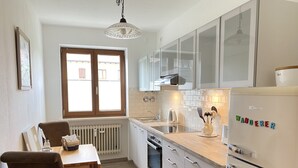 A modern kitchen with white cabinets, a built-in oven, and a refrigerator. There is a window with a view of the outside, and a dining area with a table and chairs. - Experience Chiemsee, mountains & lakes - apartment for up to 4 people, 2 bedrooms, balcony (Marquartstein)