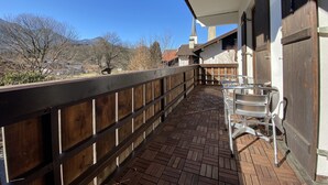 A covered balcony with a wooden railing, clear glass chairs, and a view of mountains and a church steeple. - Experience Chiemsee, mountains & lakes - apartment for up to 4 people, 2 bedrooms, balcony (Marquartstein)