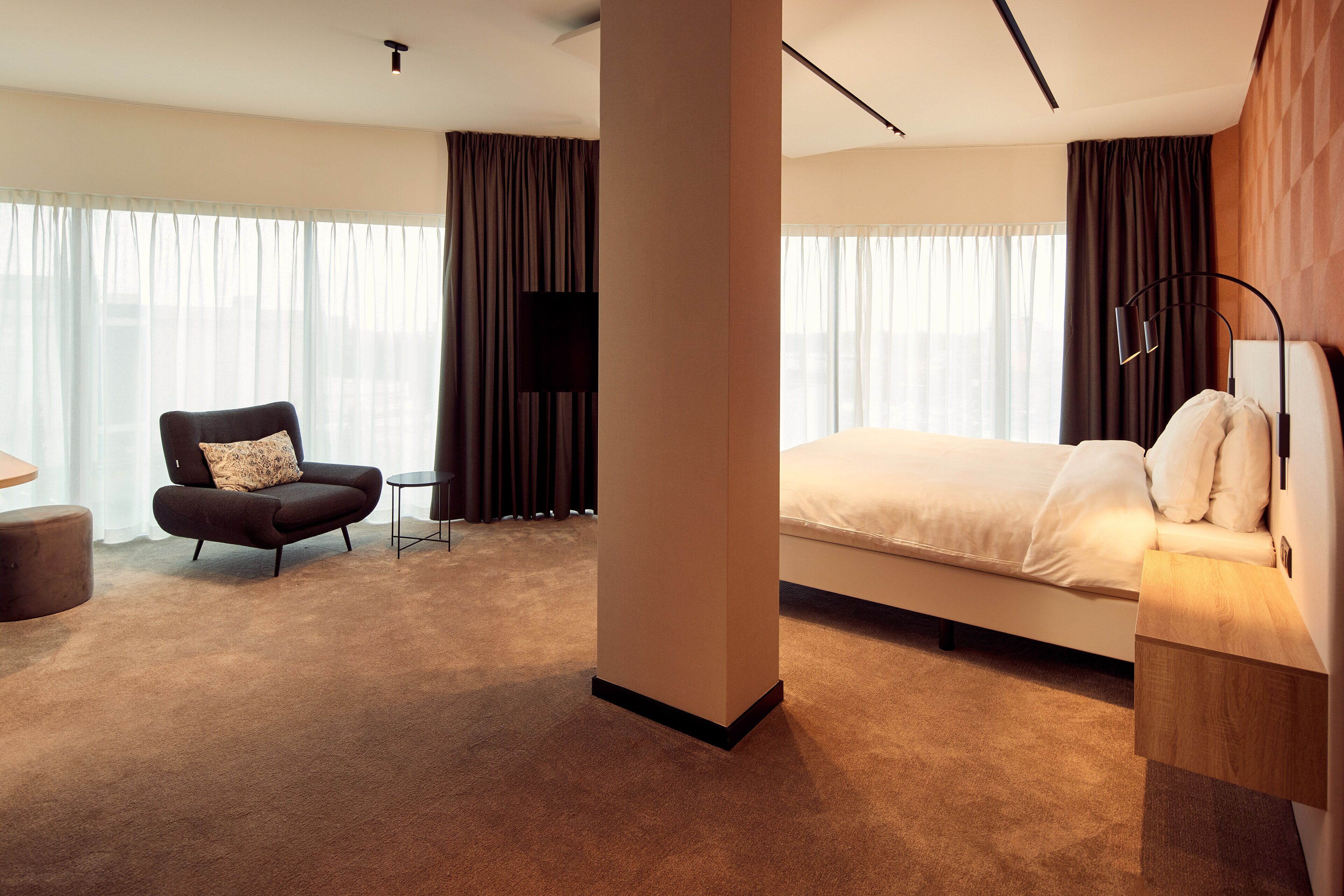 A modern hotel room with a large bed, a dark armchair, a small table, and a TV.