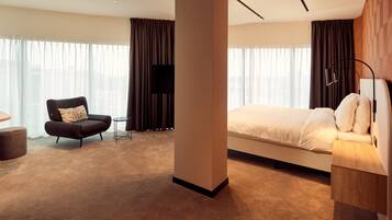 A modern hotel room with a large bed, a dark armchair, a small table, and a TV.