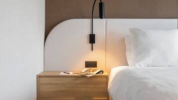 Een netjes opgemaakt bed met witte lakens, een houten nachtkastje met een lamp en een tijdschrift op het nachtkastje.