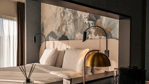 A modern hotel room with a large bed, a headboard with abstract art, two bedside lamps, a glass of water, and a coffee maker on the nightstand.