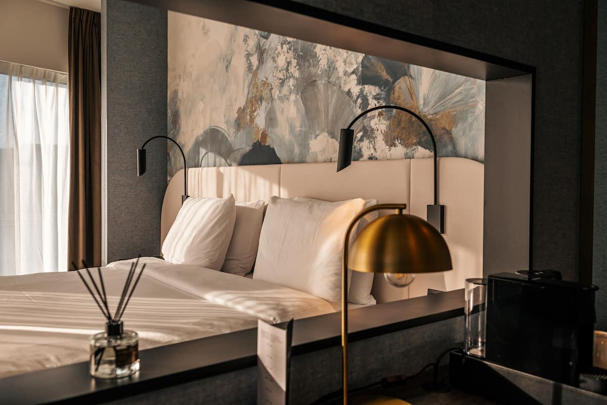 A modern hotel room with a large bed, a headboard with abstract art, two bedside lamps, a glass of water, and a coffee maker on the nightstand.