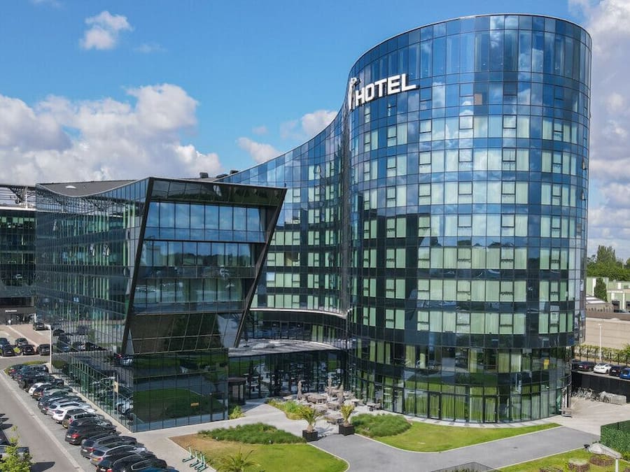 Un hotel modern amb façana de vidre corba, un pàrquing i una zona enjardinada amb bancs.