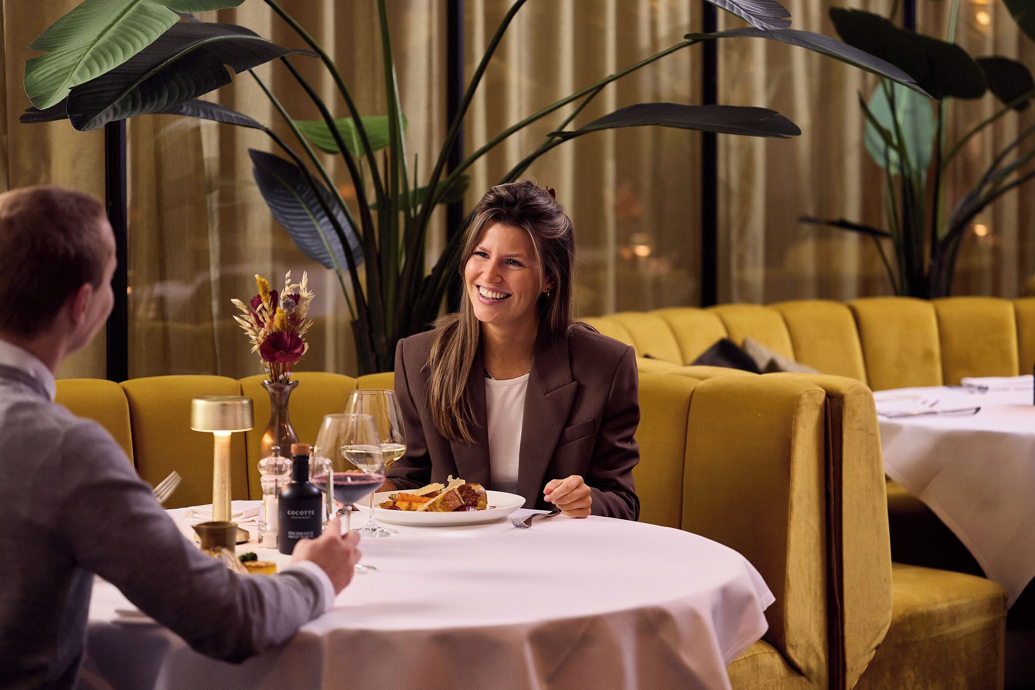 A dining table with a white tablecloth, a plate of food, and a bottle of COCOTTE wine. There are two people seated at the table, and a yellow sofa is visible in the background.