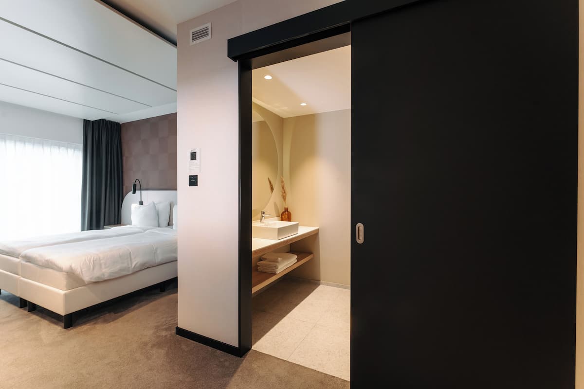 A modern hotel room with a bed, a bathroom with a sink and mirror, and a door leading to another room.