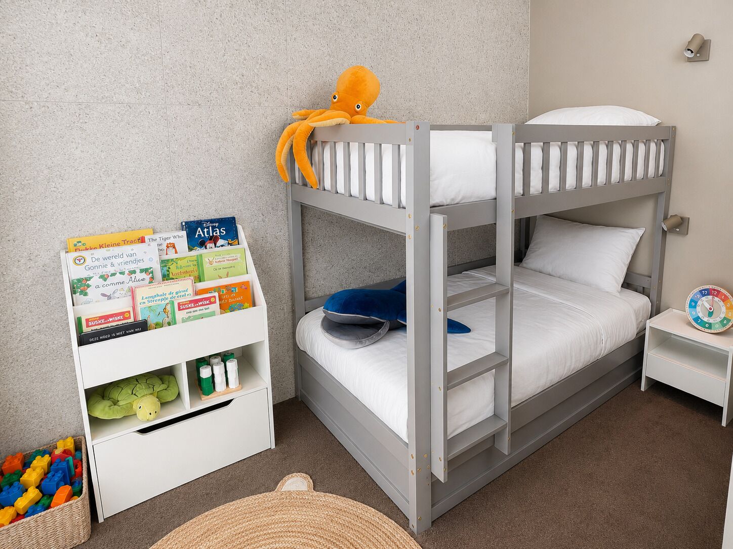 A bunk bed with a white and gray color scheme, a small bedside table with a clock, and a bookshelf with children's books and toys.
