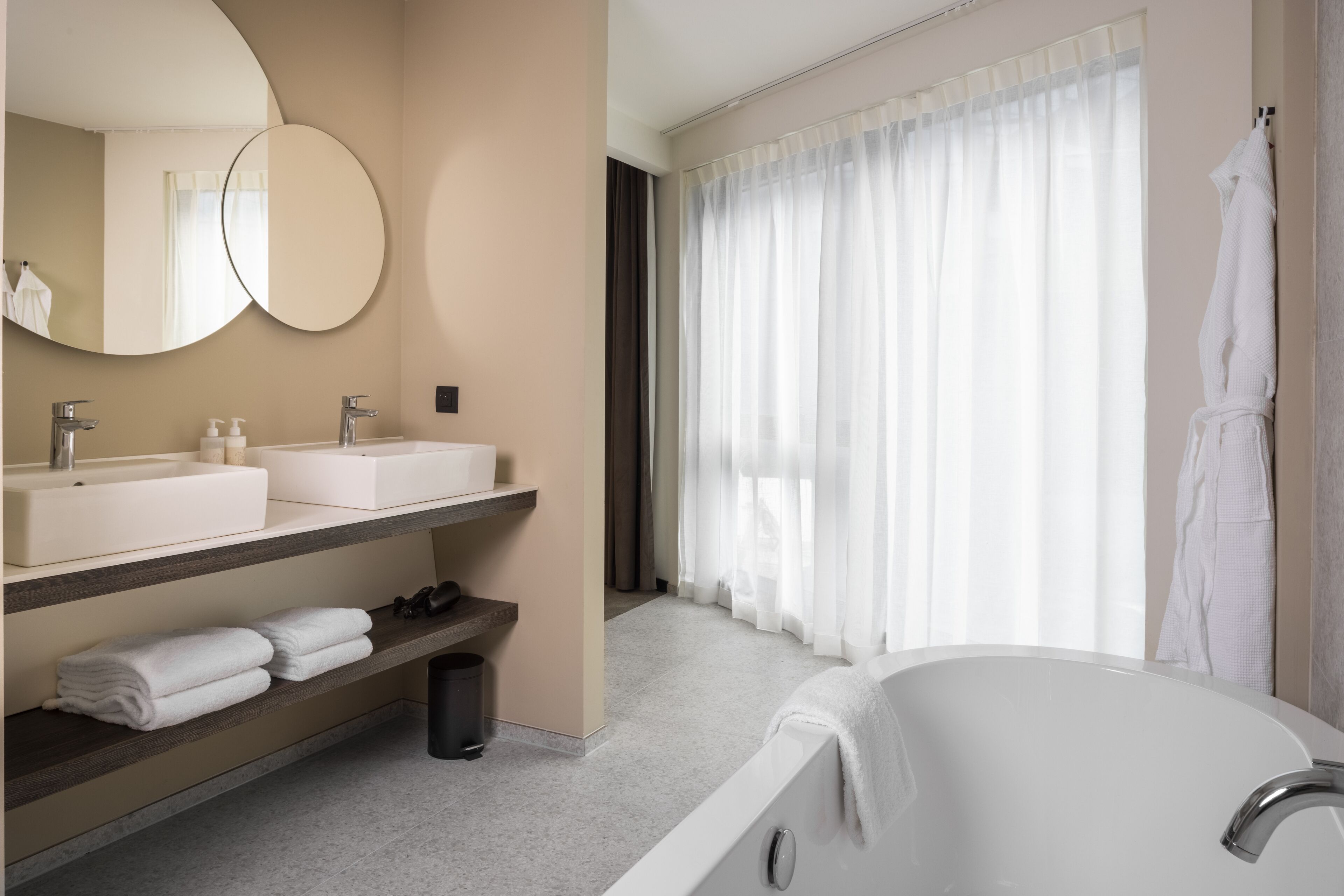 A modern bathroom with a large bathtub, a round mirror, a sink with a vanity, and a window with sheer curtains.