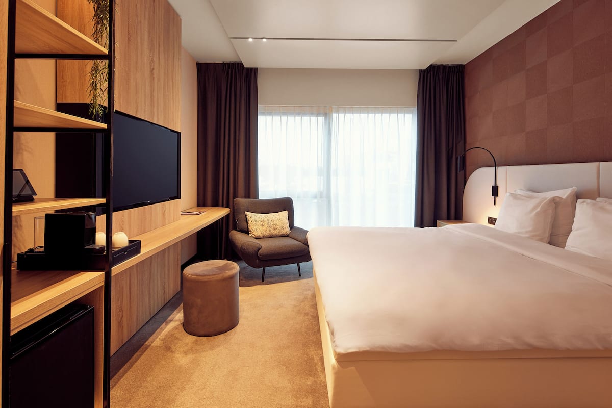 A modern hotel room with a large bed, a TV on a wooden shelf, a small seating area, and a window with curtains.
