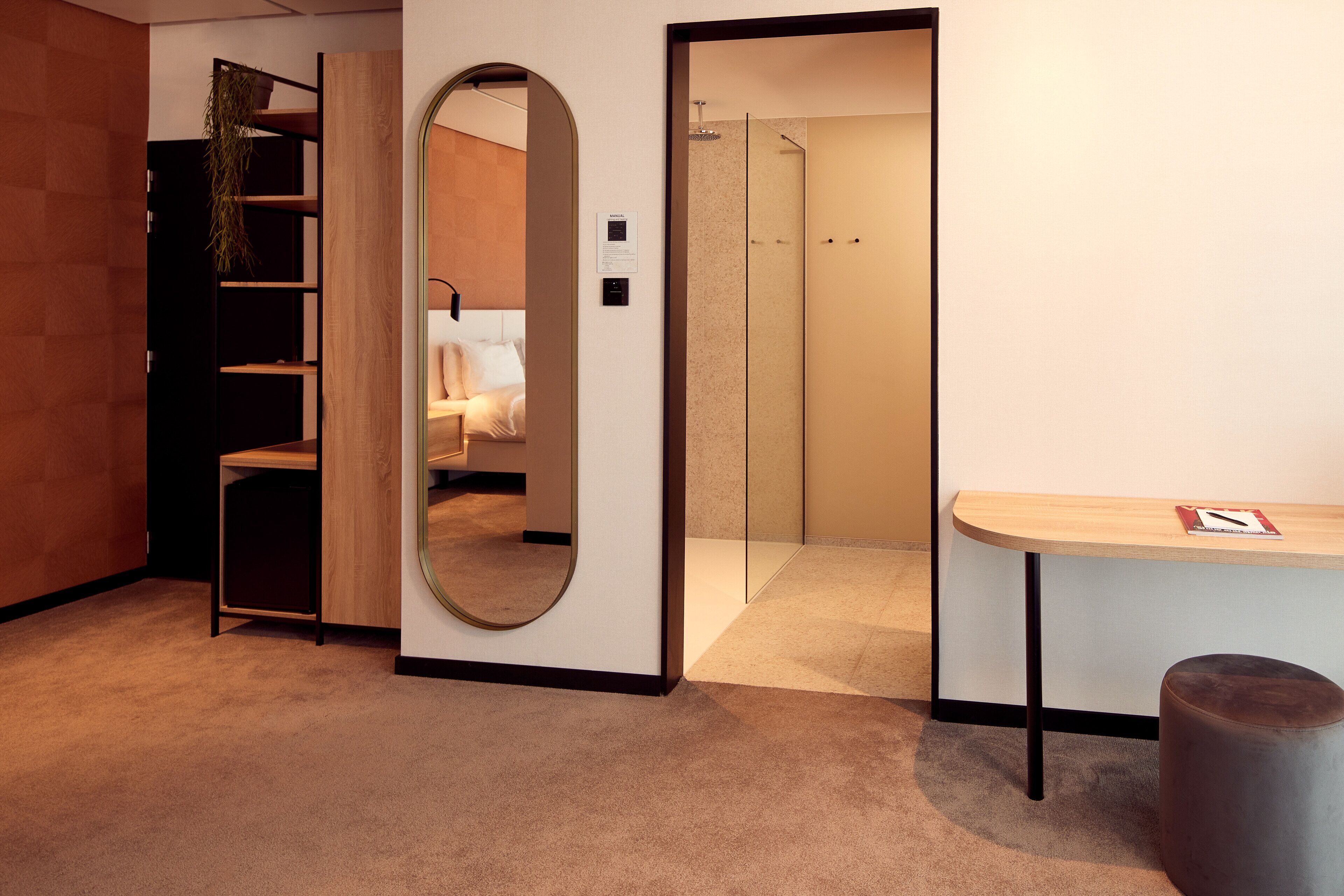 Habitación de hotel moderna con una estantería de madera, un espejo redondo, una mesita pequeña y un área de ducha.