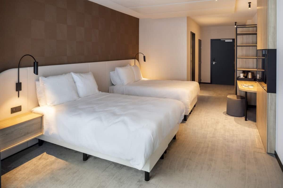 A hotel room with two beds, a bedside table, a lamp, a shelf with a cup and a book, and a door leading to another room.