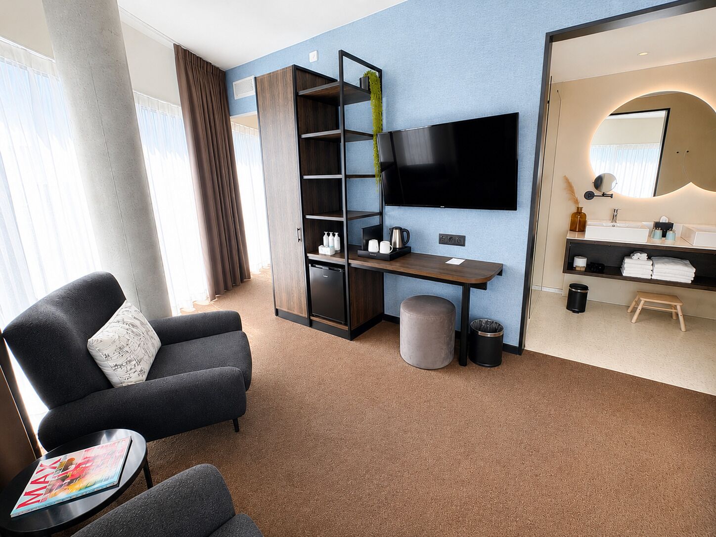 Een moderne hotelkamer met een bank, een klein tafeltje met een tijdschrift, een televisie, een bureau met een koffiezetapparaat, een prullenbak en een badkamer met een wastafel en spiegel.