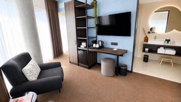 Een moderne hotelkamer met een bank, een klein tafeltje met een tijdschrift, een televisie, een bureau met een koffiezetapparaat, een prullenbak en een badkamer met een wastafel en spiegel.