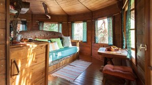 Interior rústico de una cabaña con techo de madera, una cama con cojín verde, una mesa de comedor con una cesta de fruta y una ventana con vistas al exterior.