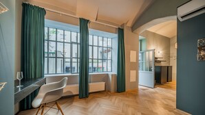 A modern hotel room with a large window, a dining area with a white chair, and a kitchen area visible through an open door. - easy-flat Neustiftgasse - AC Studio Apartments near MuseumsQuartier (Vienna)