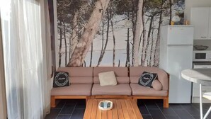 Un salon avec un canapé beige, deux coussins, une table basse en bois et un coin cuisine équipé d’un réfrigérateur et d’un four à micro-ondes.