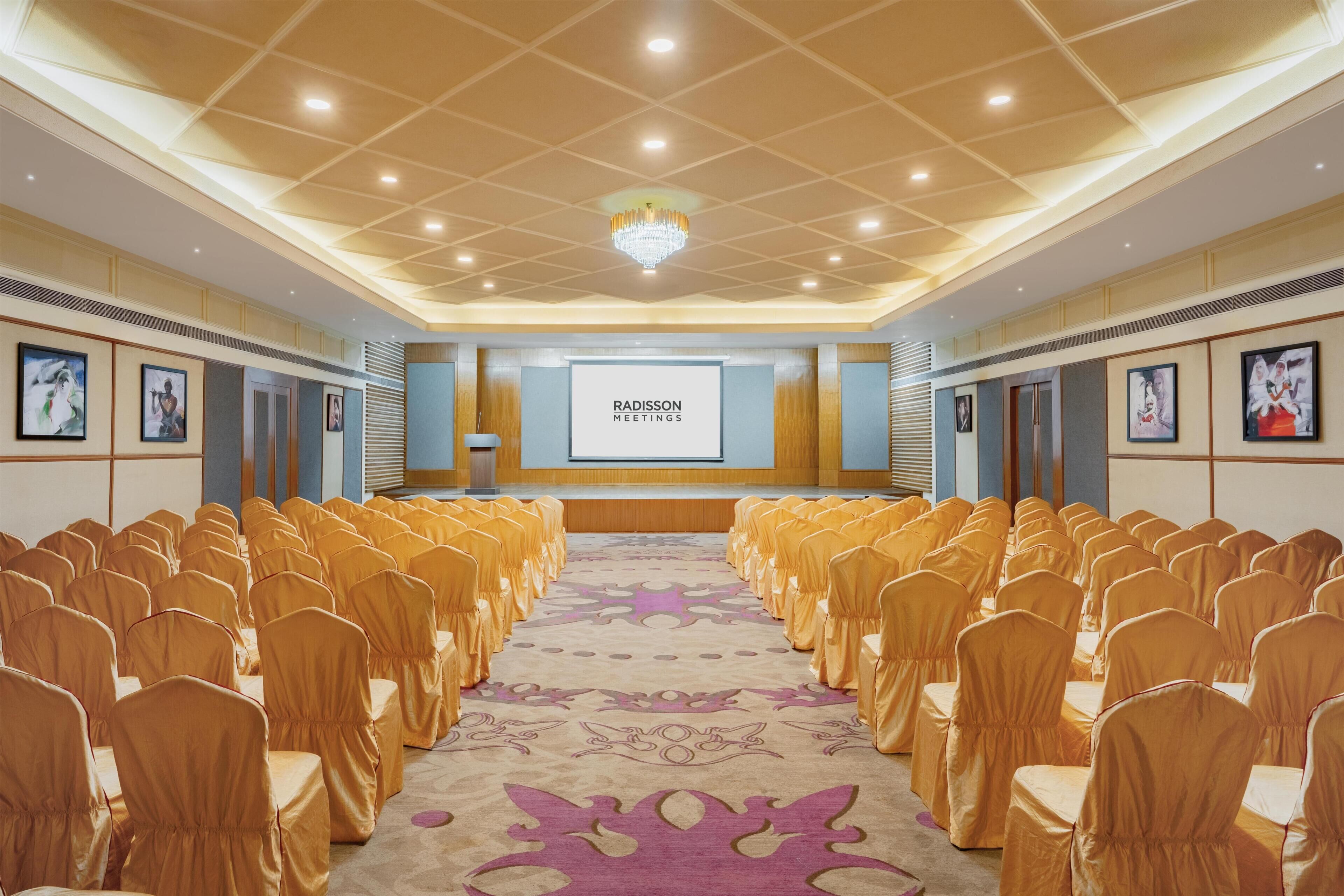 Un salón de conferencias con filas de sillas, un escenario con una pantalla y una araña.