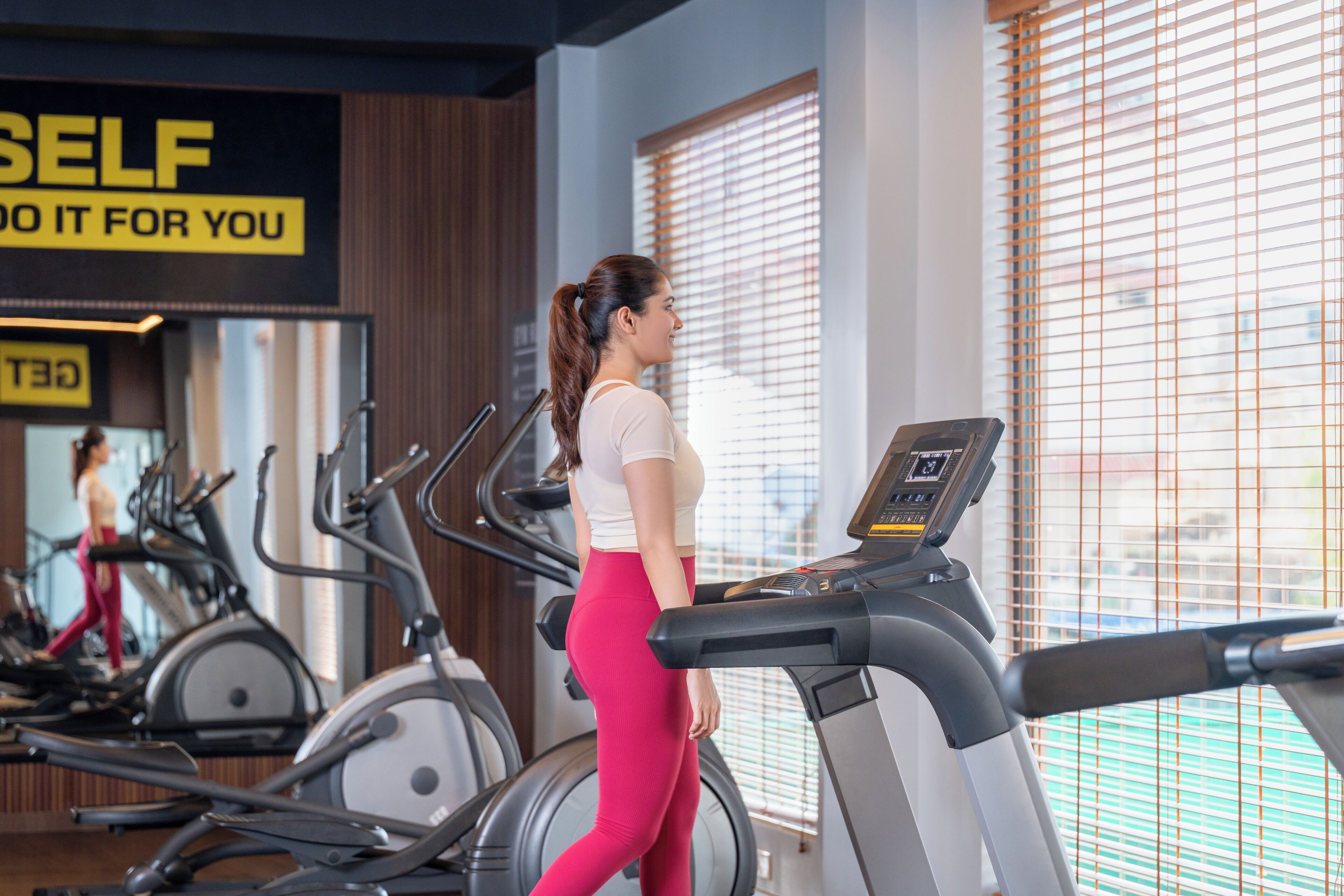 A gym with treadmills, a woman walking on one, and a mirror reflecting the room.