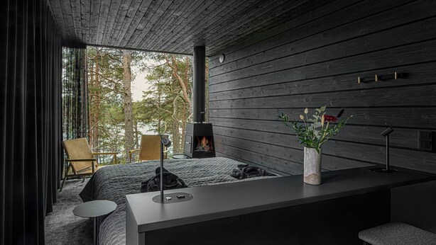 A modern, dark-themed room with a large window, a fireplace, a bed, a small table, and a bar area. - The Baro (Inga)