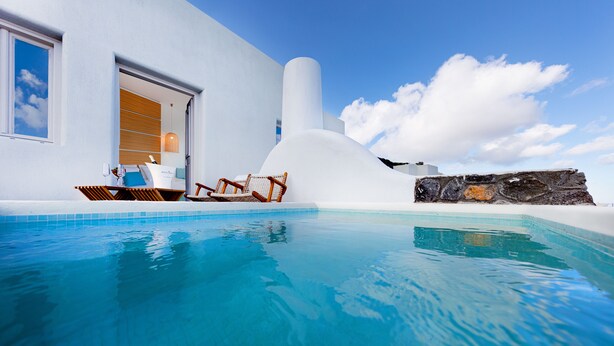 Un área de piscina moderna con agua azul cristalina, un edificio blanco y un muro de piedra.