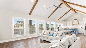 Uma sala de estar espaçosa com um amplo sofá modular cinza, teto de madeira com vigas aparentes e cozinha com armários brancos e uma ilha central.