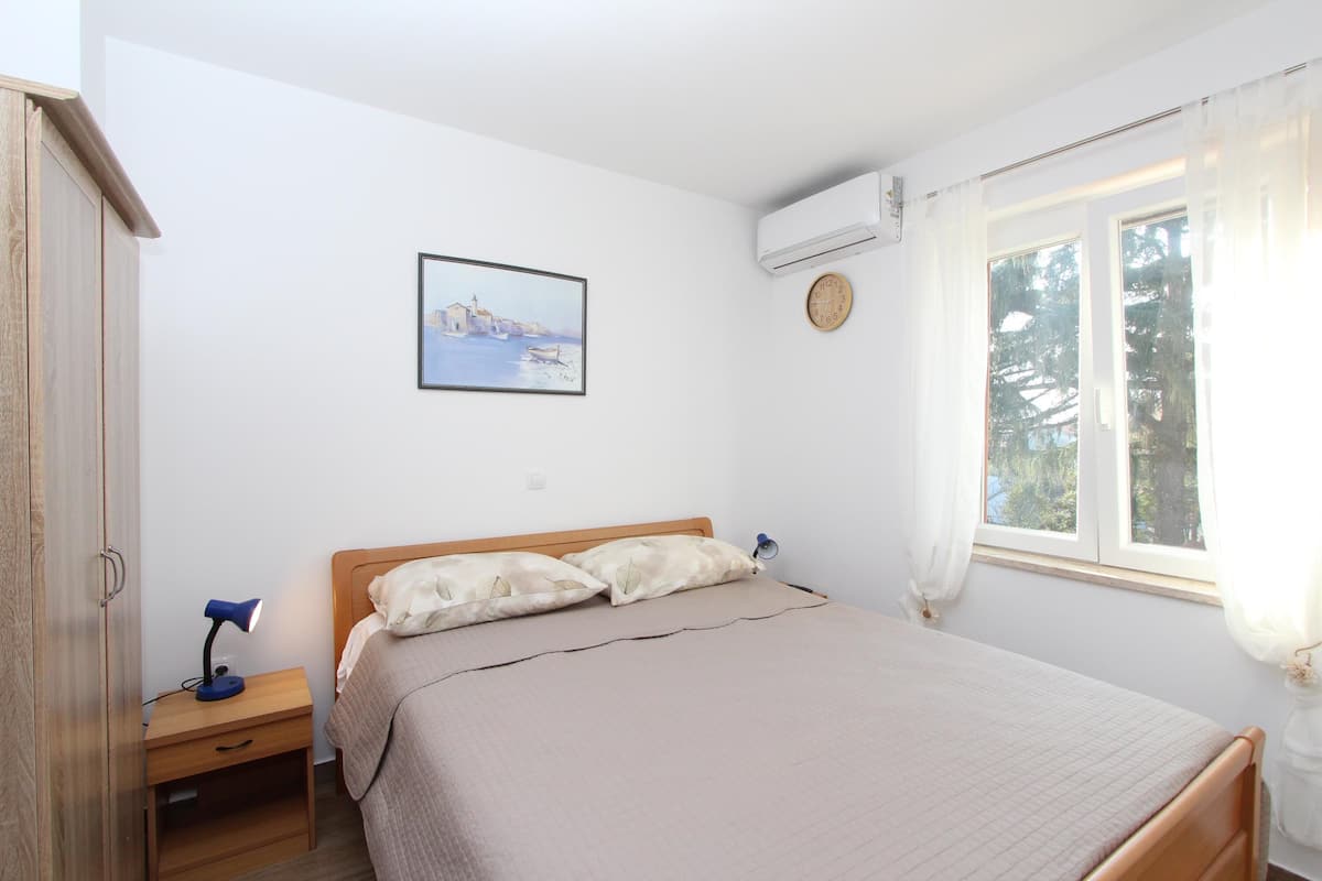 A bedroom with a wooden bed, a nightstand with a lamp, a wall clock, and a window with curtains.