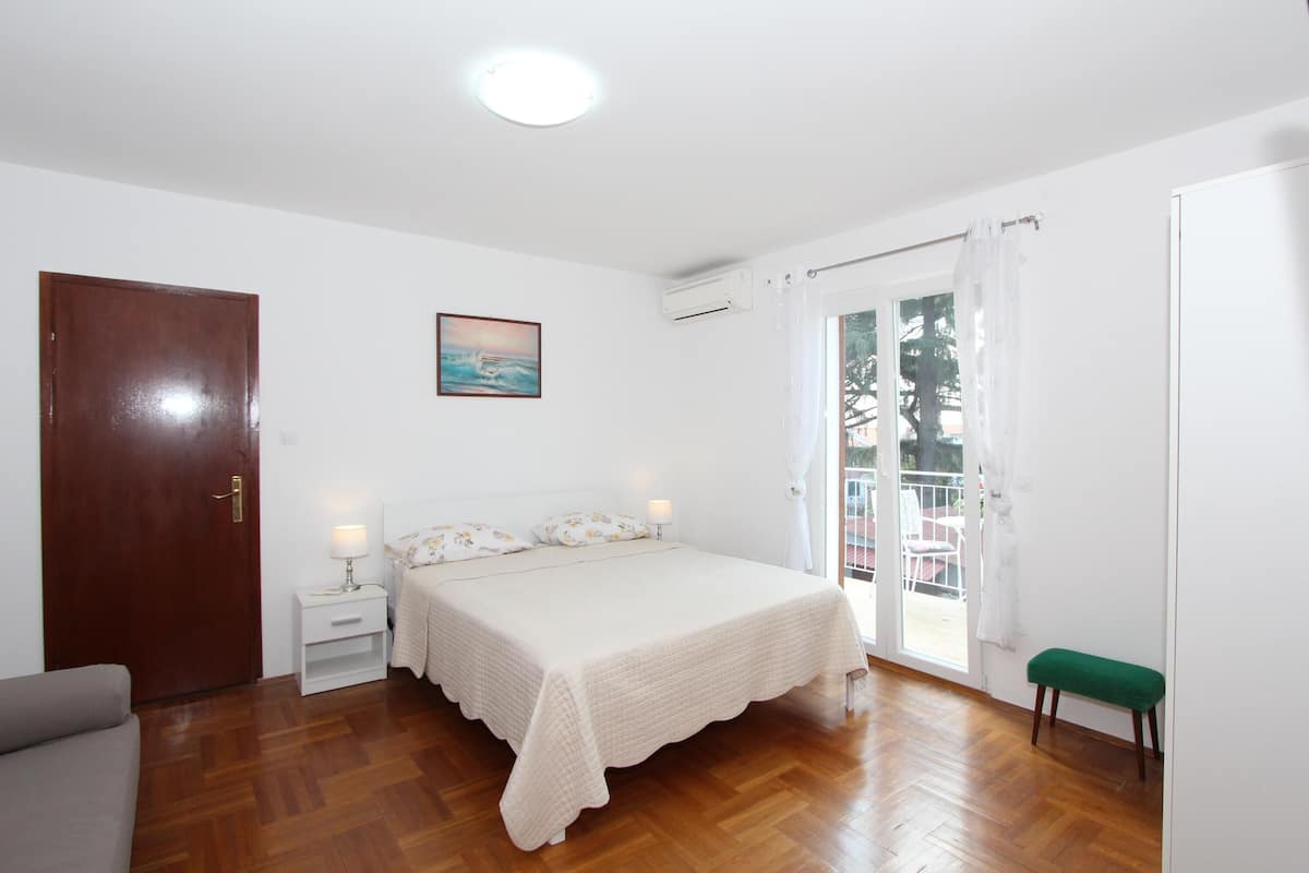 A bedroom with a wooden floor, a bed with a white bedspread, a nightstand with a lamp, a green bench, and a door.