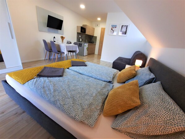 A modern bedroom with a large bed, a small sofa, and a dining area with a TV and kitchen in the background.