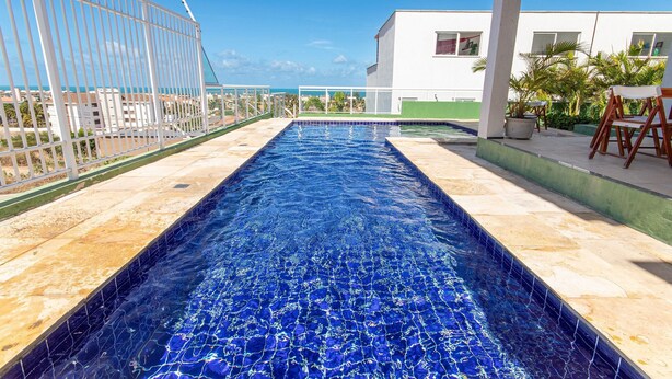 A swimming pool with a view of the sea, a white building, and a fenced area. - Triplex Hidromassagem em Porto das Dunas (Aquiraz)