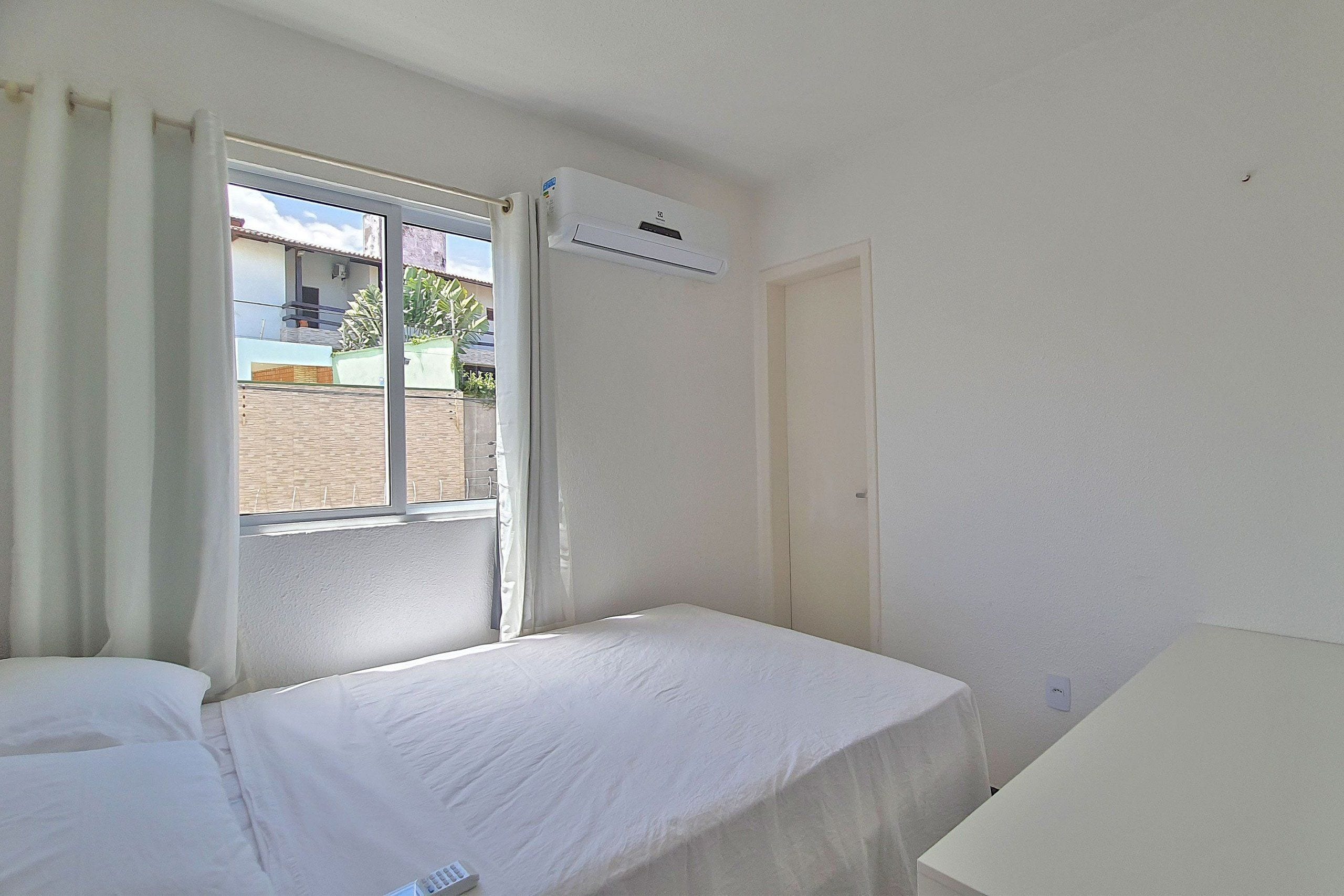 A hotel room with a bed, a window with curtains, an air conditioning unit, and a door.