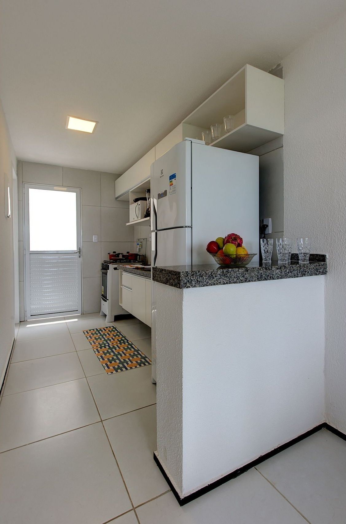 A compact kitchen with white cabinets, a refrigerator, and a countertop with a fruit bowl.