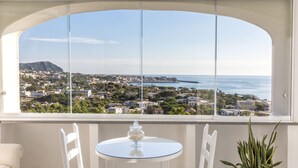 Ein Balkon mit Glastisch, weißen Stühlen, einem Blumentopf und Blick auf eine Küstenstadt.