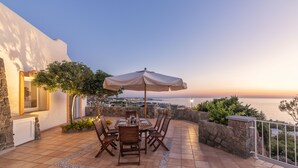Eine Terrasse mit Tisch und Stühlen, einem Sonnenschirm und Blick auf das Meer bei Sonnenuntergang.