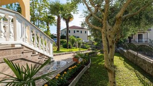 A well-maintained garden with a stone pathway, a white balustrade, and a variety of plants including yellow flowers and palm trees. - Apartment Šerbečić 4 with sea view for 4 persons on the island of Krk (Malinska)