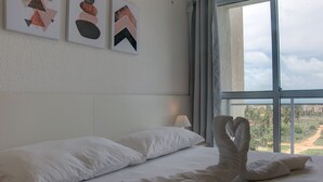 A hotel room with a large window, a bed with white linens, and abstract art on the wall. - Chalé Família em Porto das Dunas (Aquiraz)