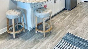 A modern kitchen with white cabinets, a stainless steel refrigerator, a microwave, and a sink. There are two bar stools and a sign that reads "BEACH" on the wall.