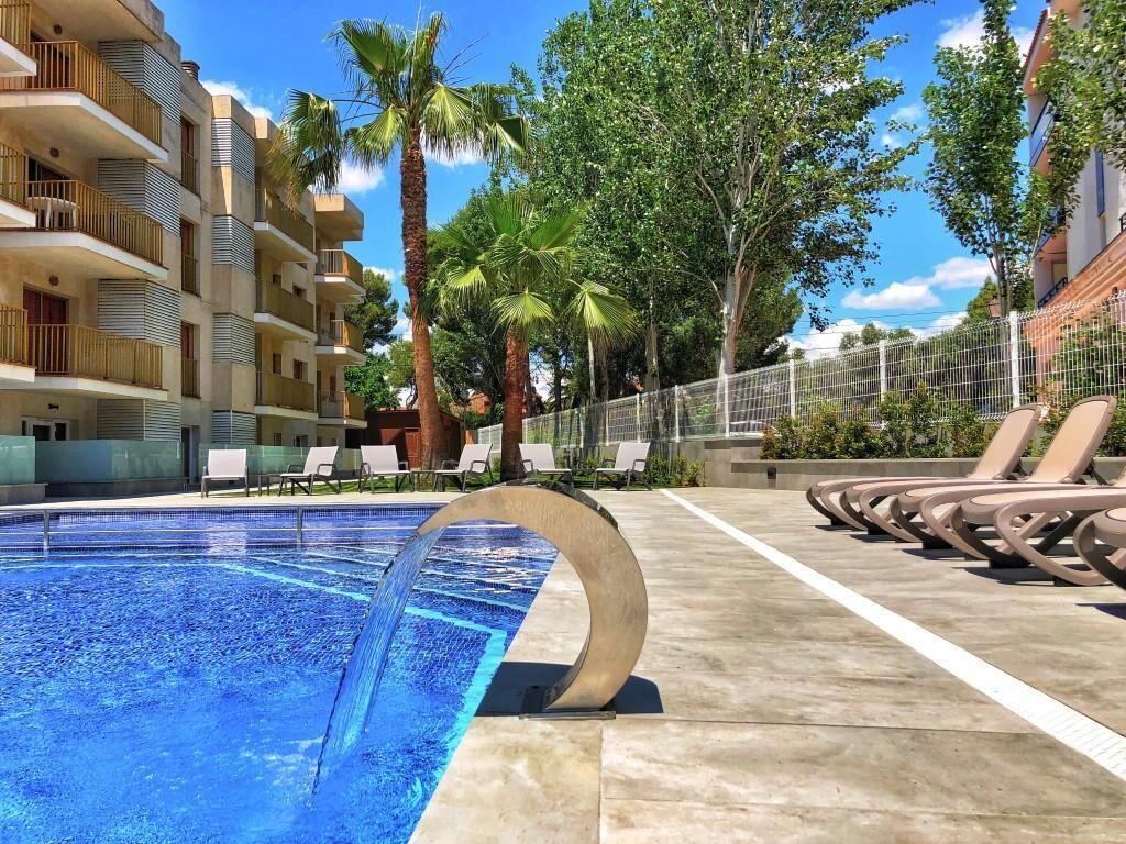 Ein Swimmingpool mit einem kreisförmigen Wasserspiel, umgeben von Liegestühlen und Palmen, mit einem mehrstöckigen Gebäude im Hintergrund.