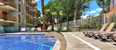 A swimming pool with a circular water feature, surrounded by lounge chairs and palm trees, with a multi-story building in the background.