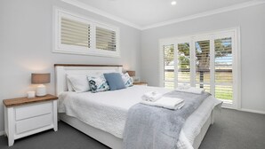 A bedroom with a large bed, two bedside tables, a nightstand, a window with blinds, and a view of greenery outside. - The Bunkhouse - quality, contemporary home (Exeter)
