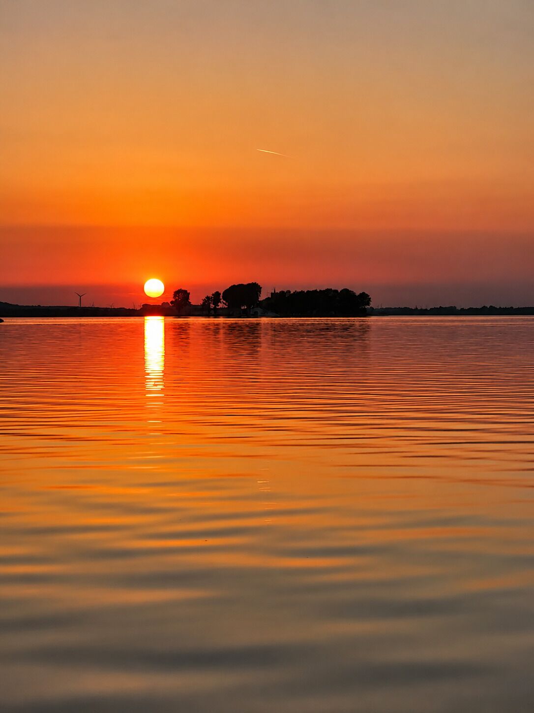 Ein Sonnenuntergang über ruhigem Gewässer mit der Silhouette von Bäumen und einer Windkraftanlage in der Ferne.
