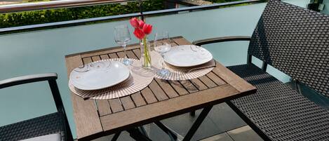 An outdoor dining area with a table set for two, chairs, and a view of a garden and building in the background.