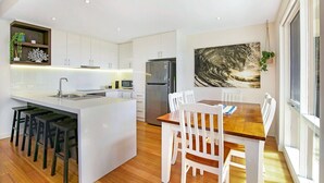 Une cuisine moderne avec des meubles blancs, un îlot central doté de tabourets de bar, un réfrigérateur en acier inoxydable et un coin repas avec une table en bois et des chaises blanches.